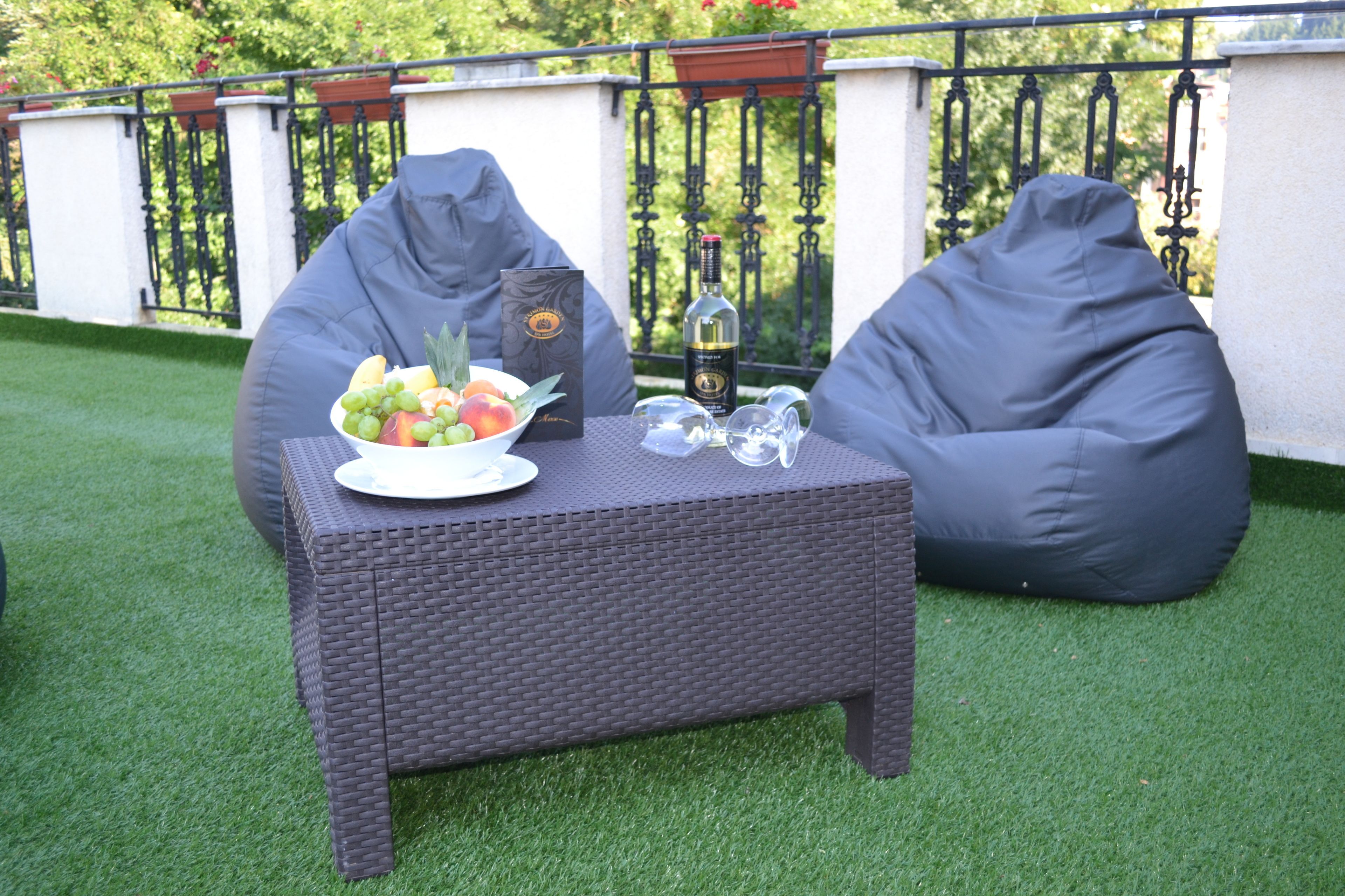 A wicker table with a bowl of fruit, a bottle, and glasses on a patio with artificial grass and a metal fence in the background.