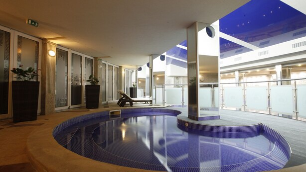 Een binnenzwembad met een blauwe betegelde vloer, omringd door een gang met glazen deuren en een plafond met een geometrisch patroon.