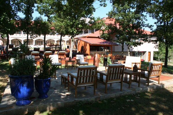 An outdoor seating area with wooden chairs and tables, surrounded by potted plants and a building with a red-tiled roof. - Hotel Mellow (Pismenovo)