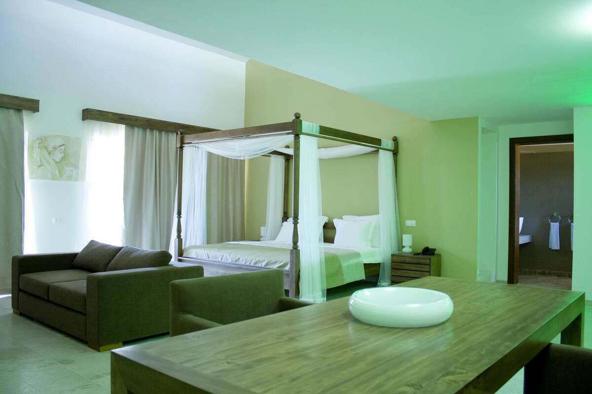 A hotel room with a four-poster bed, a sofa, a wooden table with a white bowl, and a bathroom visible through an open door.
