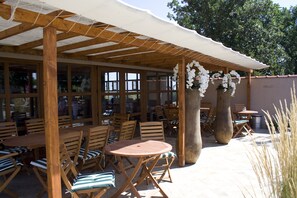 Un’area salotto all’aperto con tavoli e sedie in legno, una pergola con tenda bianca e fiori bianchi pendenti dalla struttura.