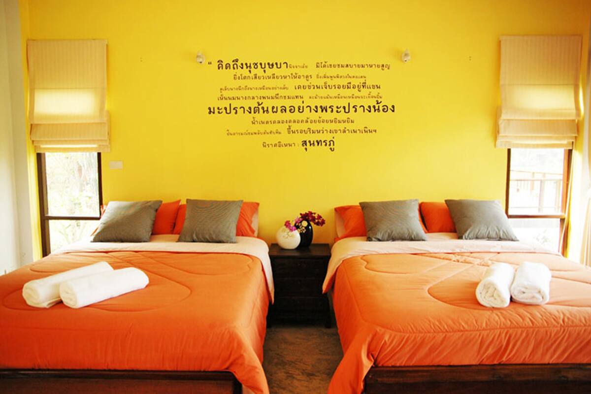 A hotel room with two beds, each with orange bedding and pillows, a small table with a vase of flowers, and a wall with Thai text.