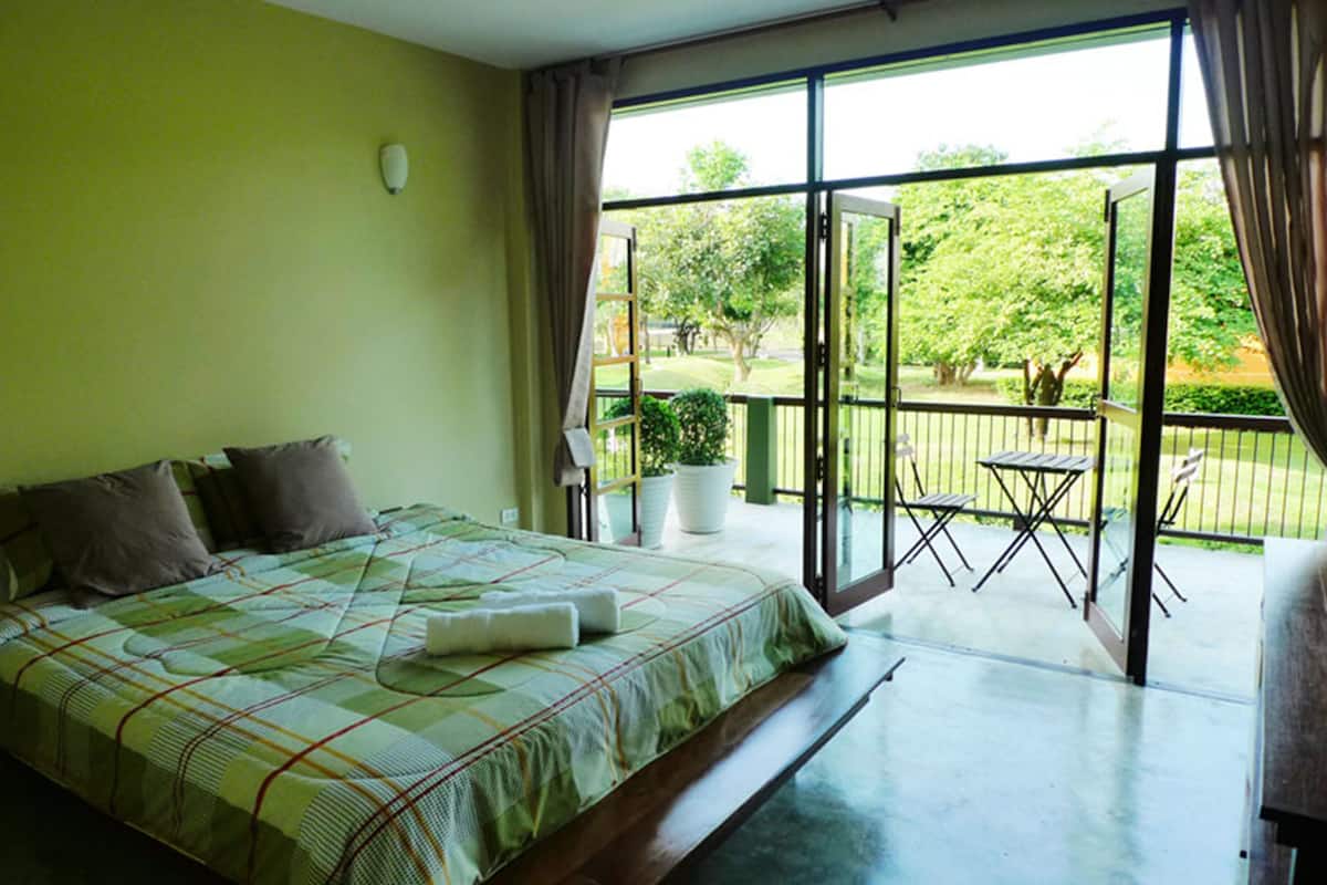 A bedroom with a large bed, a balcony with a table and chairs, and a view of greenery outside.