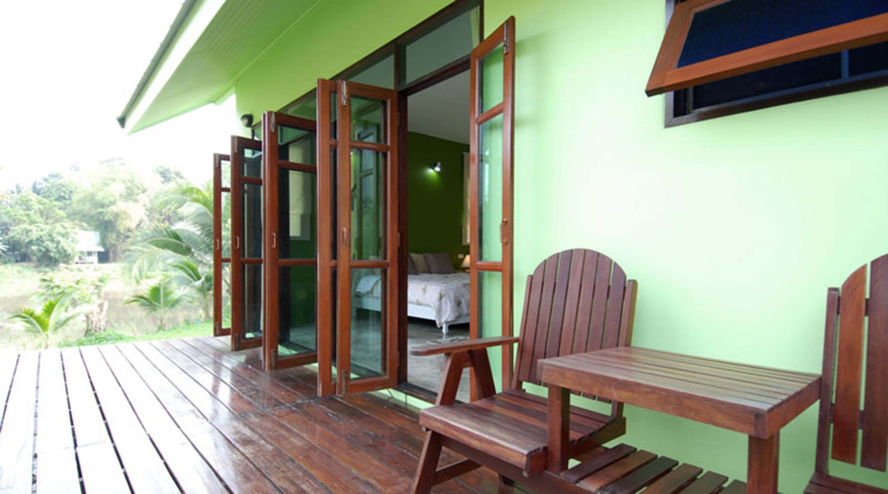 Een houten terras met een stoel en een tafel, een slaapkamer met een bed en nachtkastje, en uitzicht op groen.