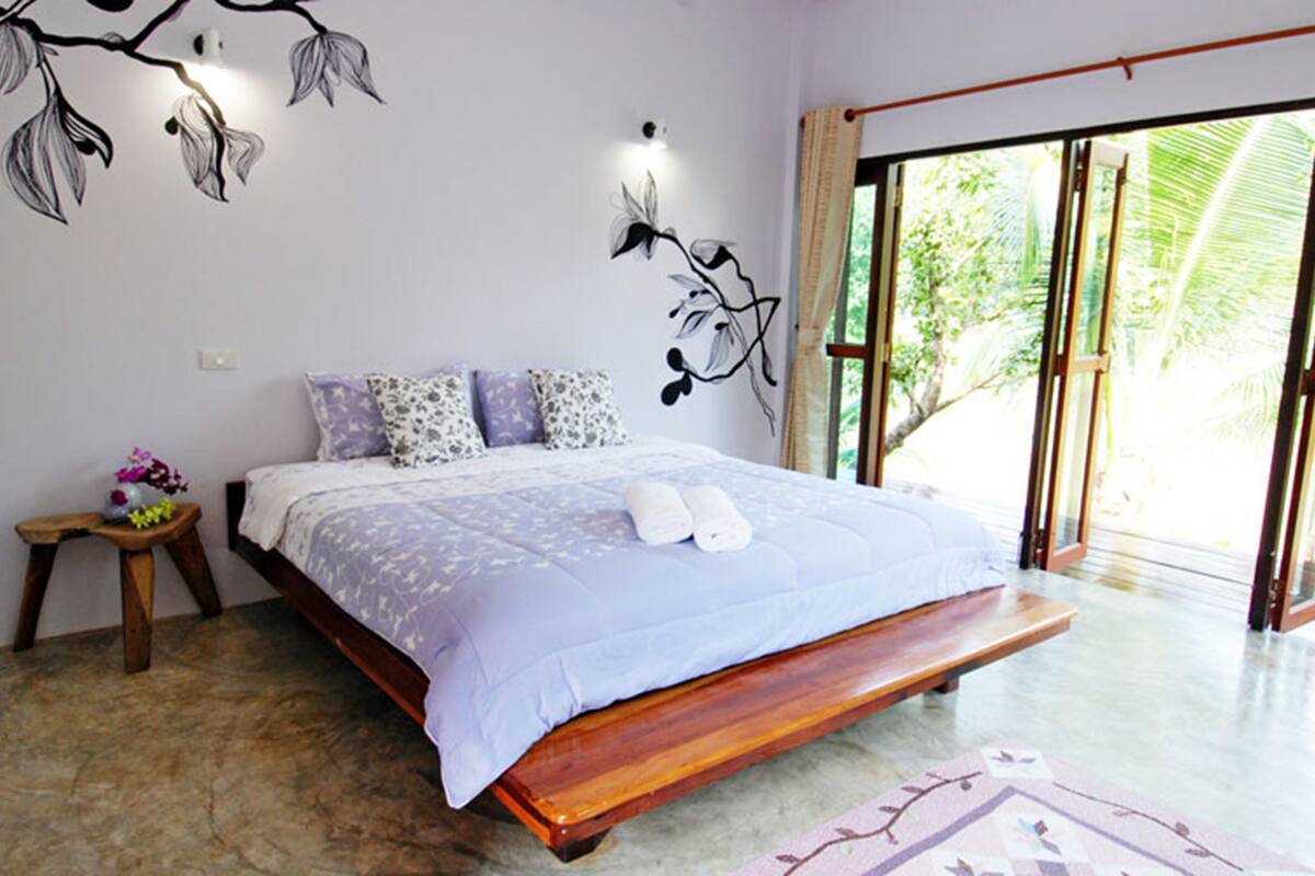 A bedroom with a bed, pillows, a nightstand, and a view of greenery through a glass door.