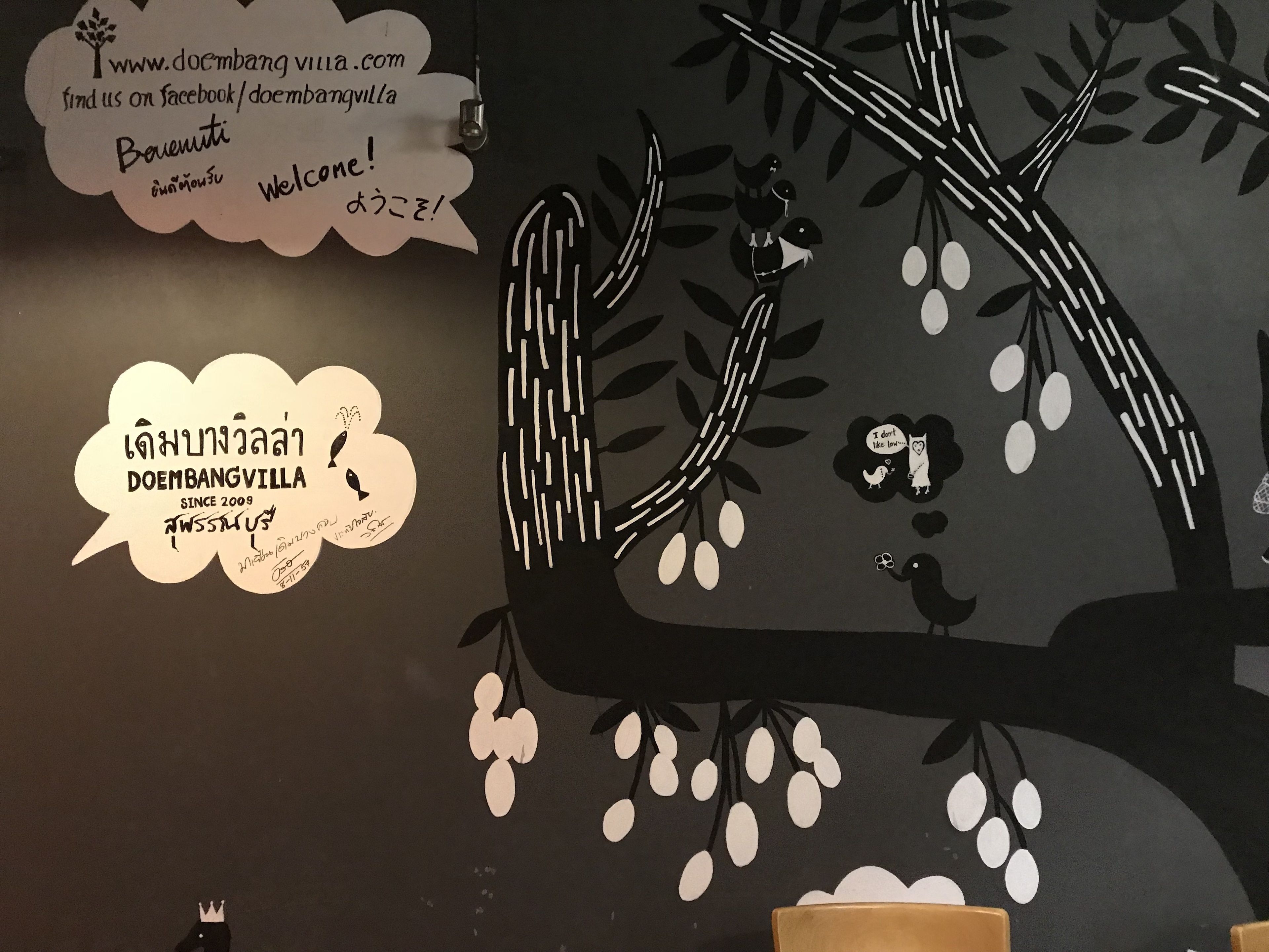 Una pared con diseño de árbol y texto en varios idiomas dando la bienvenida a visitantes en Doembangvilla.