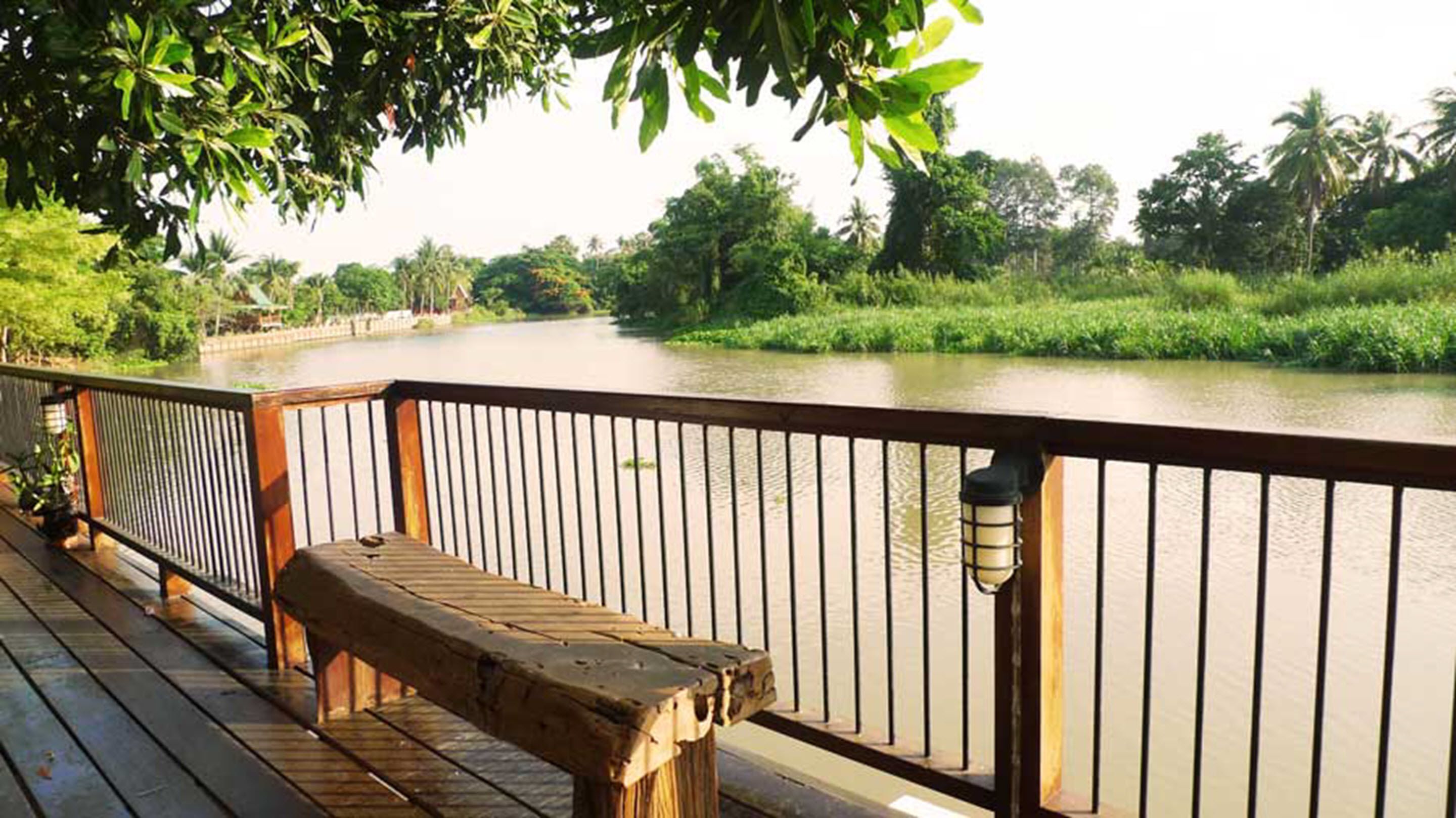 A wooden bench on a deck overlooking a calm river, with a metal railing and a lamp post.