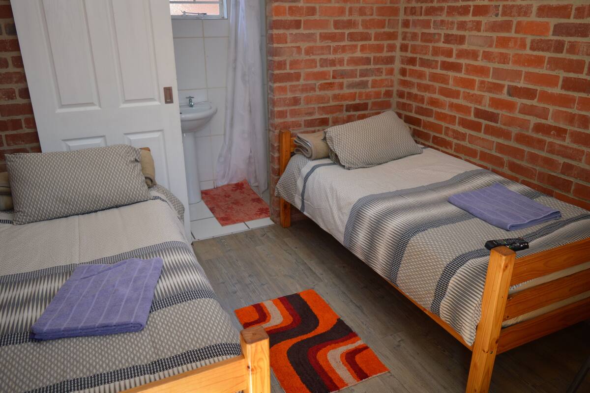 Een knusse kamer met twee eenpersoonsbedden, een houten deur, een witte wastafel en een bakstenen muur.