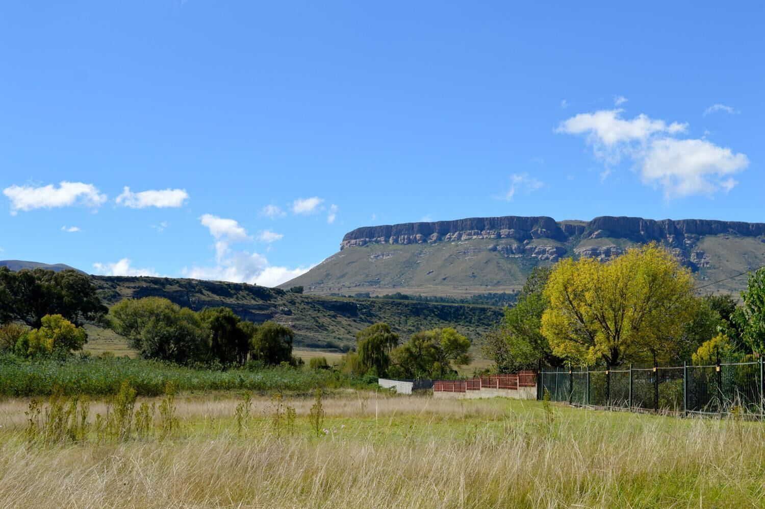 Paisagem rural com uma montanha imponente ao fundo, área cercada com um edifício e diversidade de vegetação.