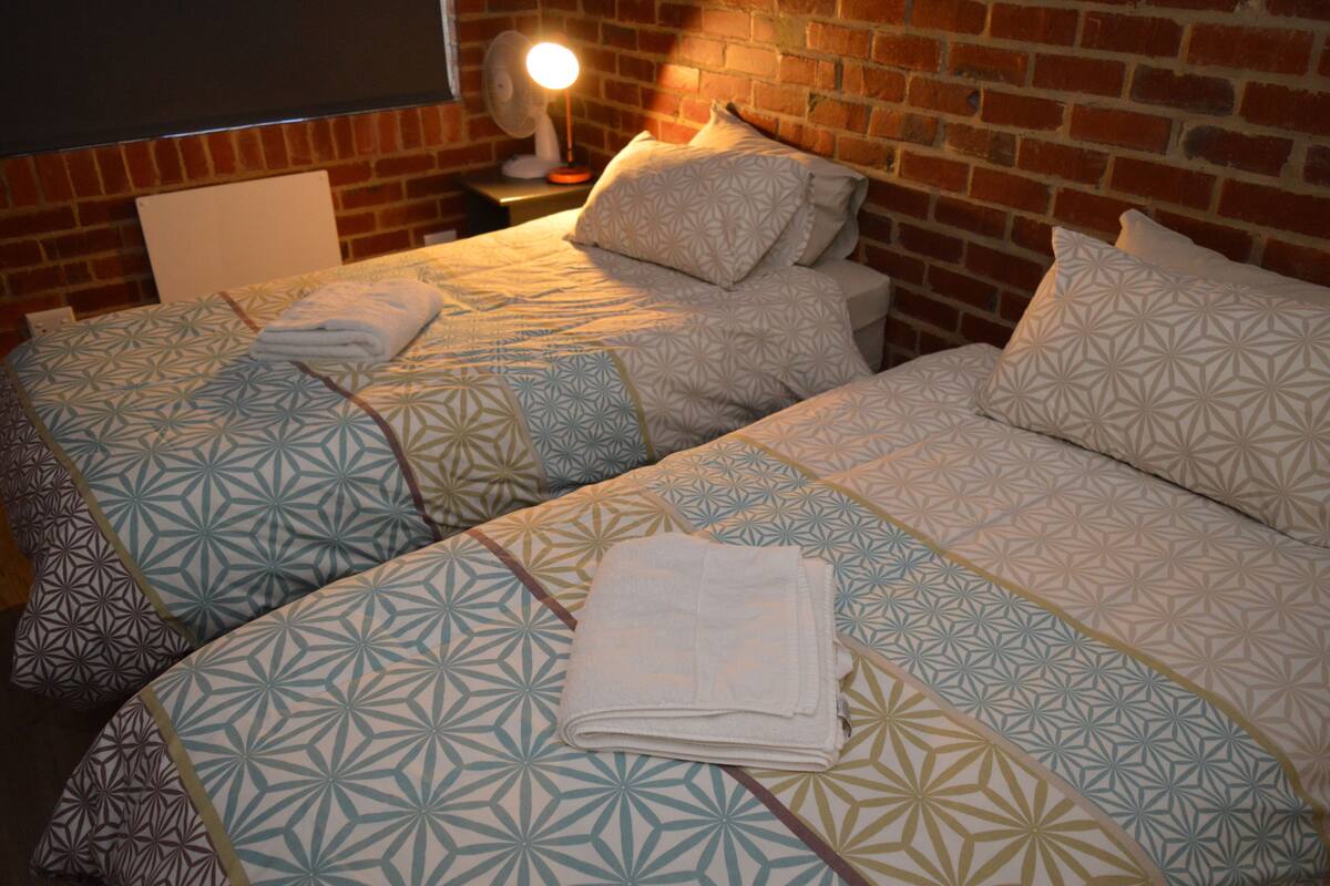 Twee bedden met beddengoed met een patroon, een nachtkastje met een lamp, en een bakstenen muur.