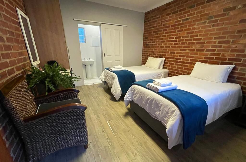 Habitación de hotel con dos camas, piso de madera, pared de ladrillo, puerta blanca y un pequeño rincón de descanso con una planta.