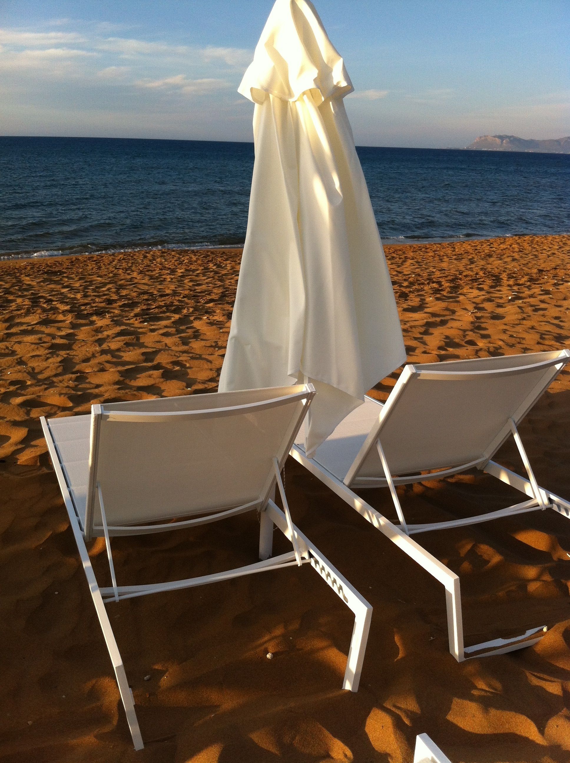 Dos sillones blancos en una playa arenosa con una sombrilla blanca, mirando al mar.