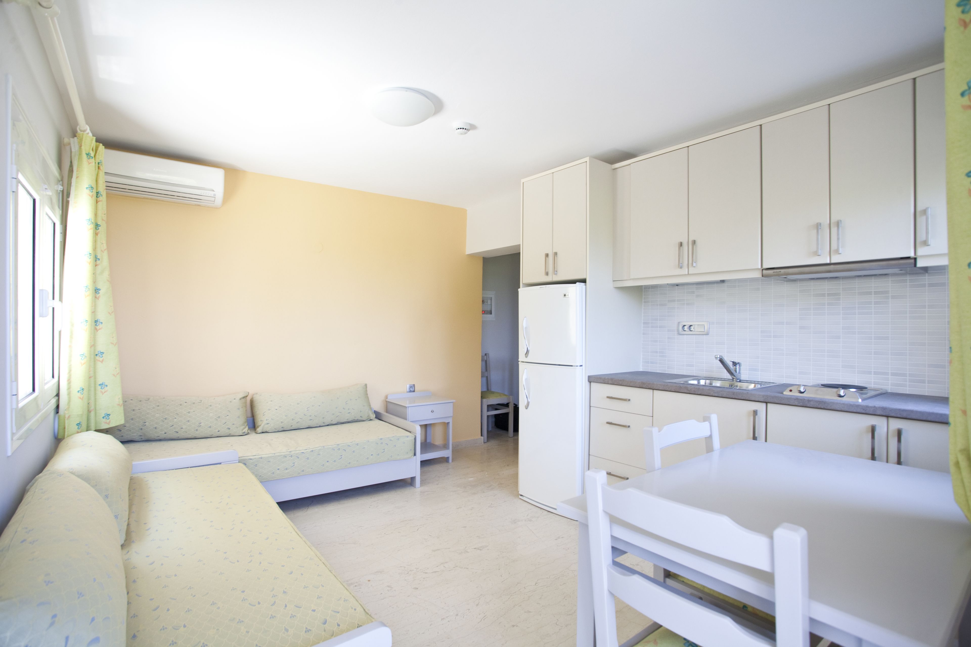 Cocina compacta con gabinetes blancos, un refrigerador y un lavamanos. Hay un pequeño comedor con una silla. Un sofá cama está apoyado contra la pared, y se ve una ventana con cortinas.
