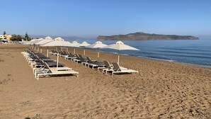 A beach with rows of sun loungers and umbrellas, a clear sky, and a distant island. - Phaedra Hotel (Chania)