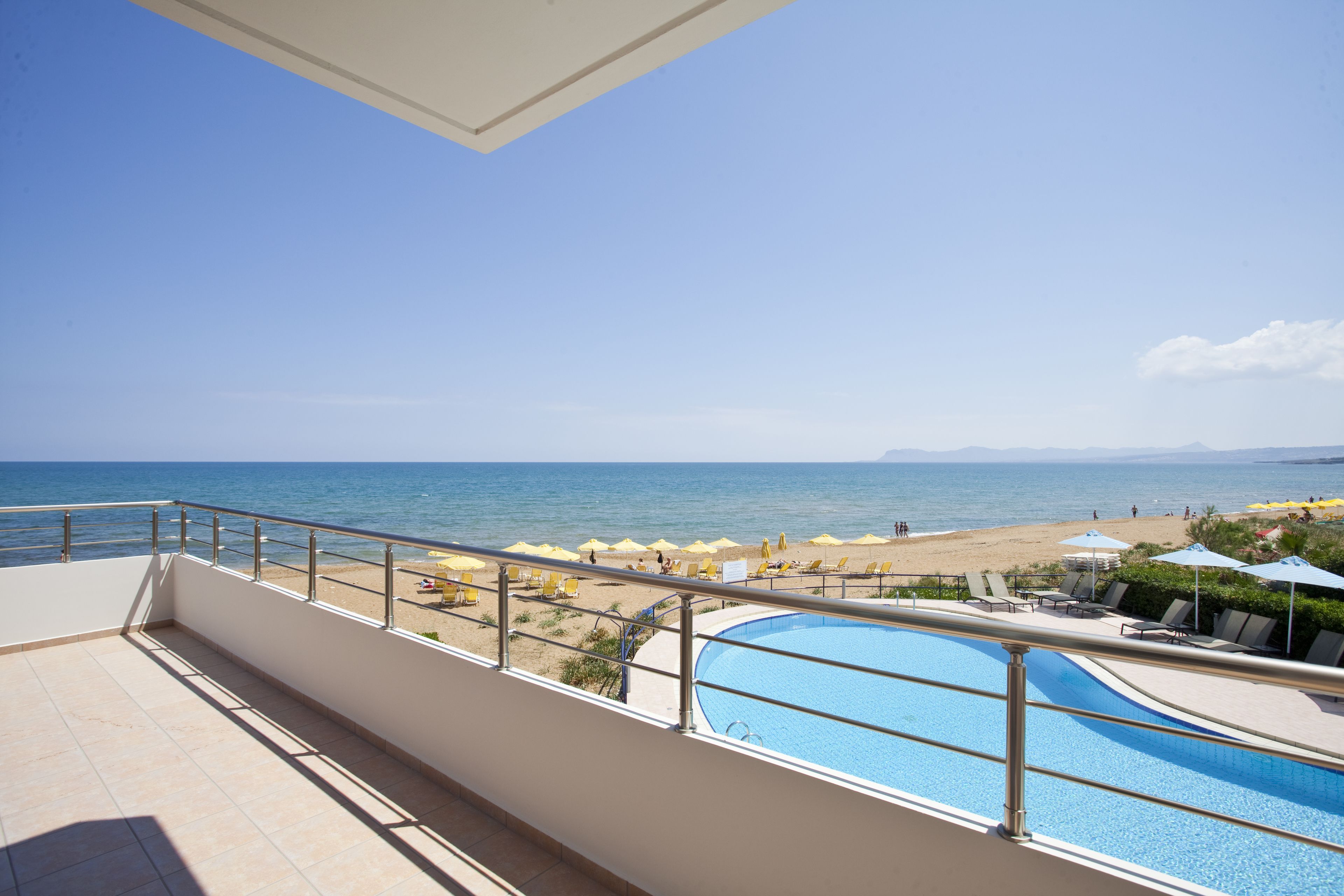 A balcony with a view of a beach, a swimming pool, and lounge chairs.
