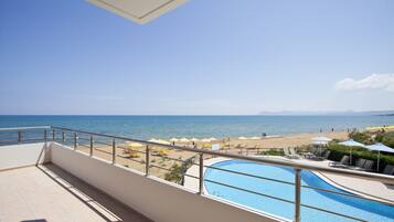 A balcony with a view of a beach, a swimming pool, and lounge chairs.
