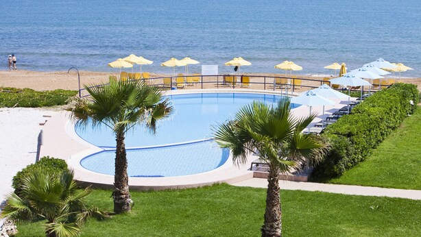 A pool with a curved edge, surrounded by palm trees and yellow umbrellas, with a beach and ocean in the background. - Phaedra Hotel (Chania)