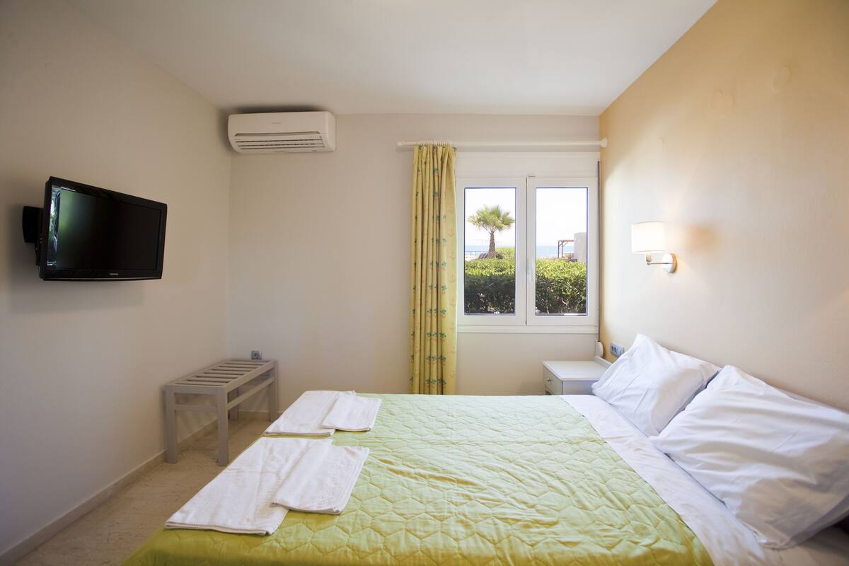 Habitación de hotel con cama, televisor montado en la pared, ventana con cortinas y una mesita con toallas.