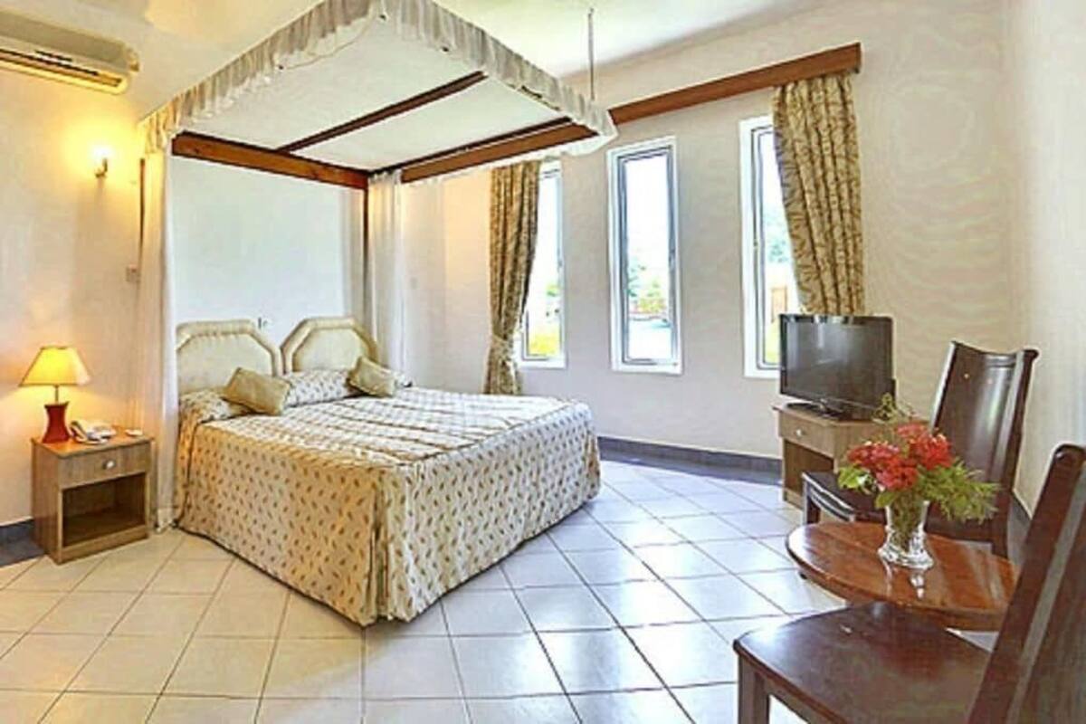 A room with a four-poster bed, a television, a dining table with chairs, and a vase with flowers.