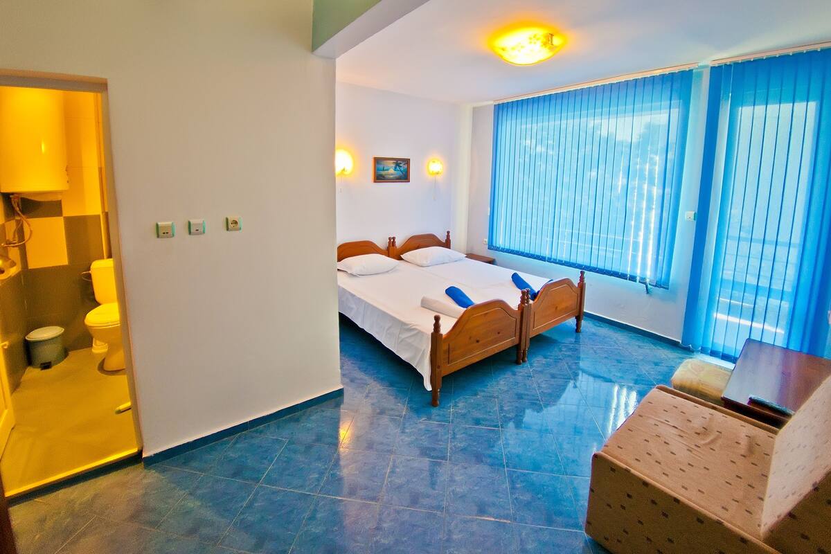 A hotel room with a bed, a window with blue curtains, a wooden chair, and a small table.