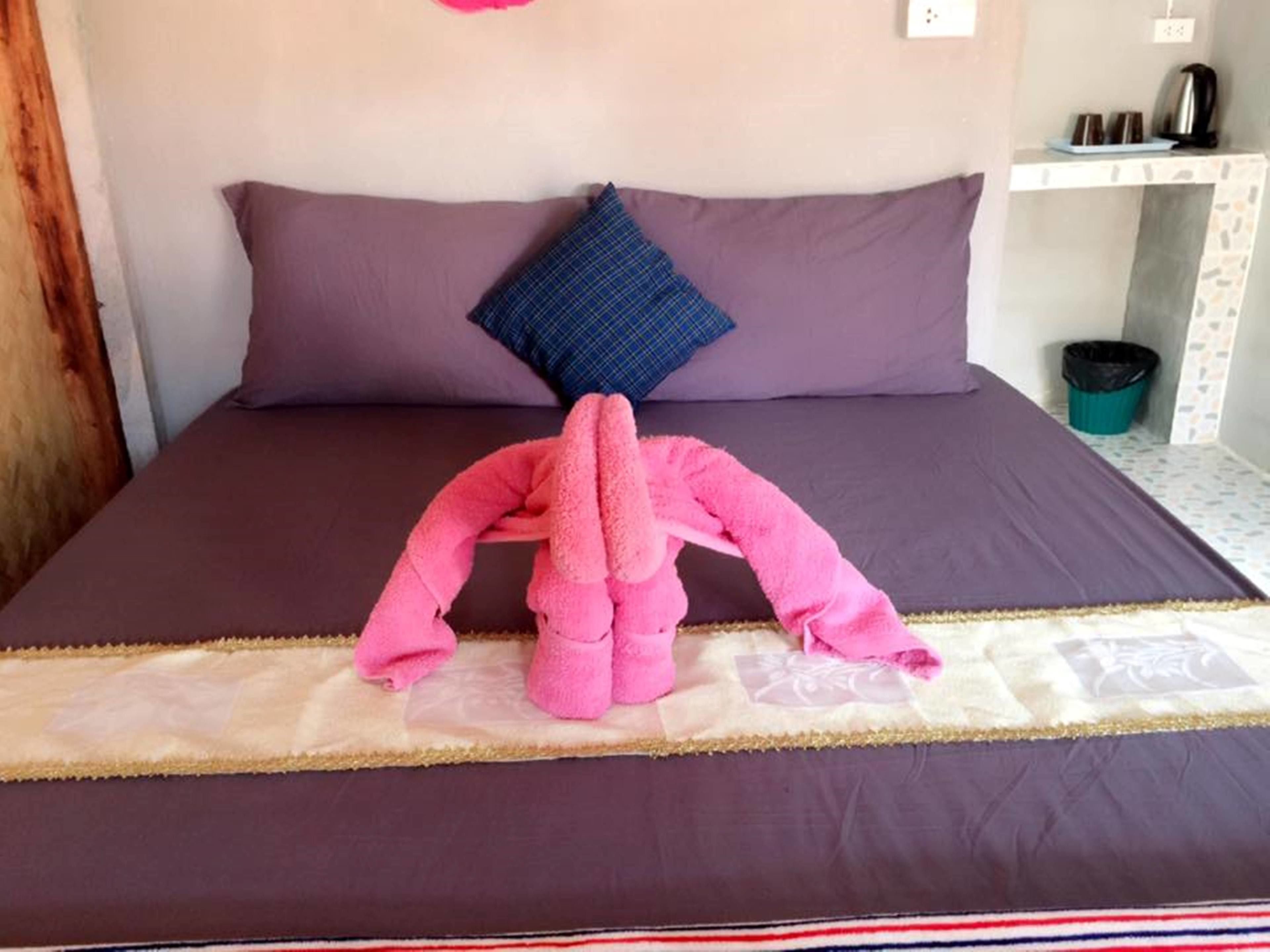 A bed with a pink towel sculpture, a purple pillow, and a small table with cups and a kettle.
