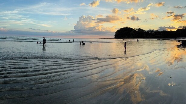 Escena de playa con gente caminando por la orilla, agua tranquila y un cielo al atardecer.