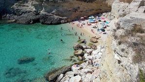 A coastal beach with clear turquoise water, rocky cliffs, and umbrellas. - Antico Camino (Muro Leccese)