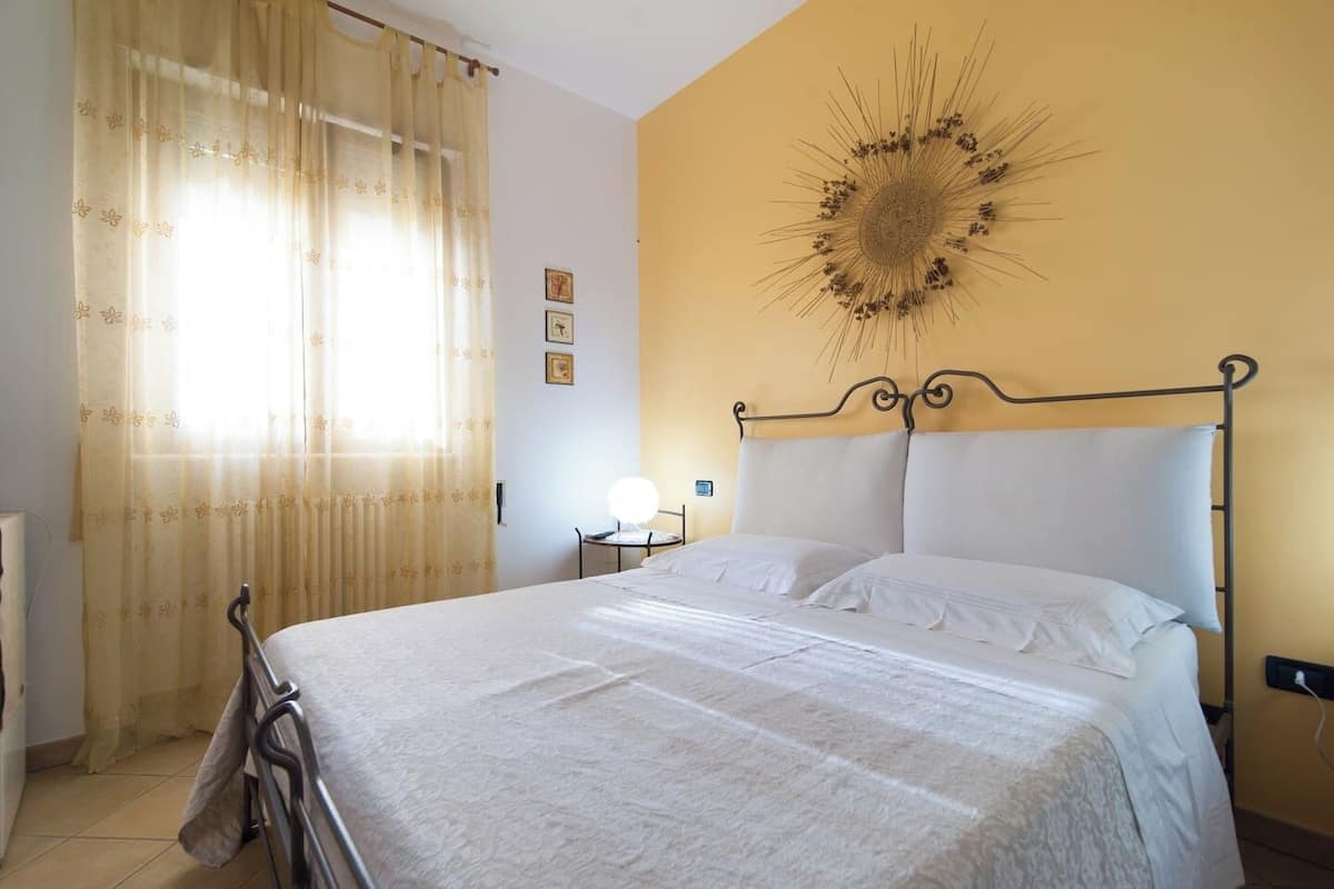 A bedroom with a metal bed frame, white bedding, a wall-mounted light, and a decorative sunburst piece.