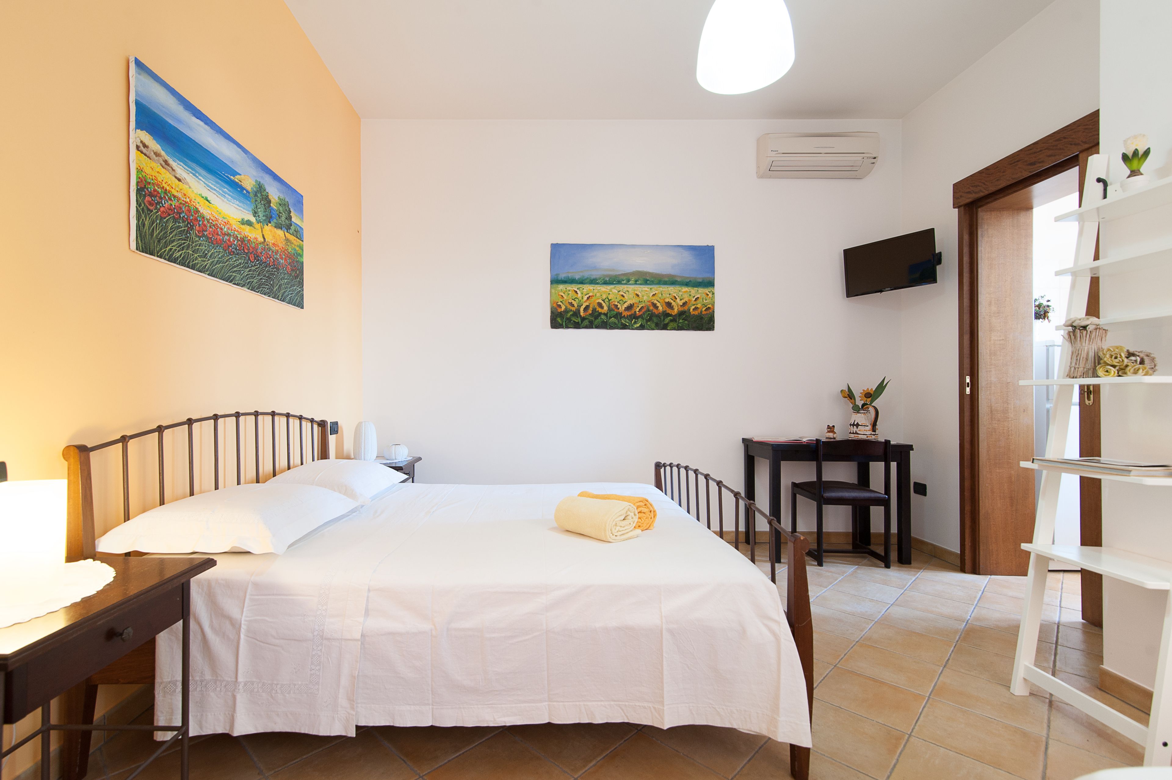 A bedroom with a bed, a desk, a chair, a TV, and two paintings on the wall.