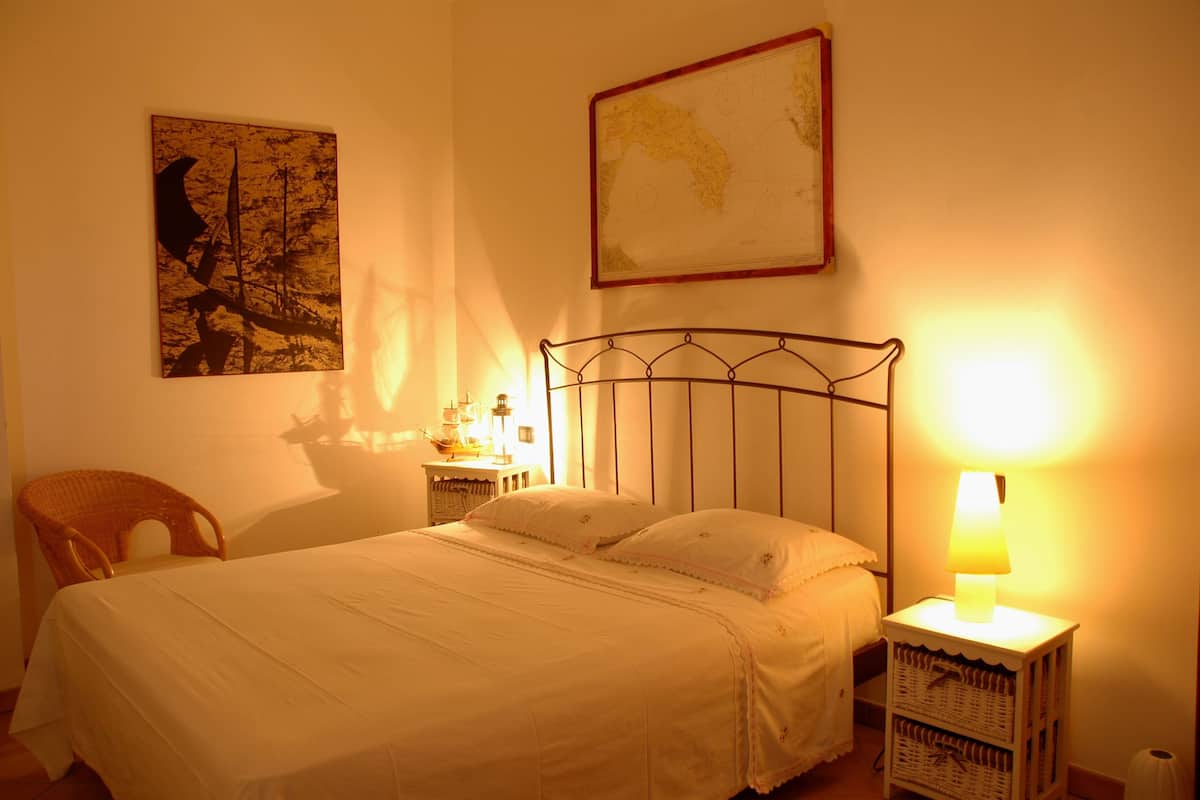 A neatly made bed with a metal headboard, a wicker chair, a nightstand with a lamp, and a map on the wall.