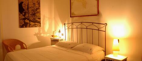 Ein ordentlich bezogenes Bett mit Metallkopfteil, ein Korbstuhl, ein Nachttisch mit Lampe und eine Karte an der Wand.