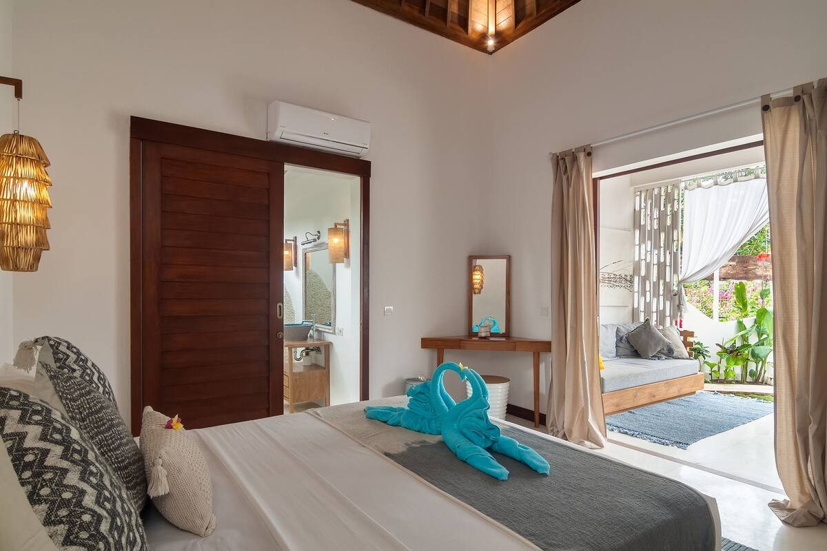 A hotel room with a bed, a wooden door, a bathroom, a small table with a mirror, and a view of the outside.