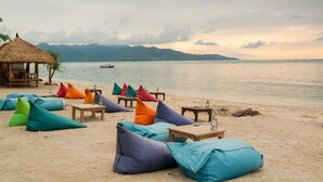 Un paisaje de playa con coloridos sillones de saco, mesas de madera, una estructura de techo de paja y un mar tranquilo con un barco a lo lejos.