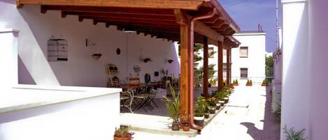 Un área de estar al aire libre techada con una pérgola de madera, plantas en macetas y una mesa de comedor con sillas.
