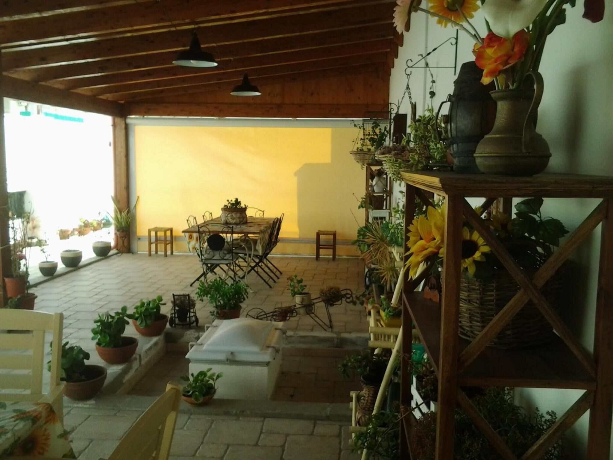 An indoor patio with a wooden table and chairs, surrounded by potted plants and a variety of flowers.