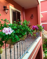 Un balcón con plantas y flores en macetas, una barandilla de metal y un edificio con fachada y contraventanas rojas.