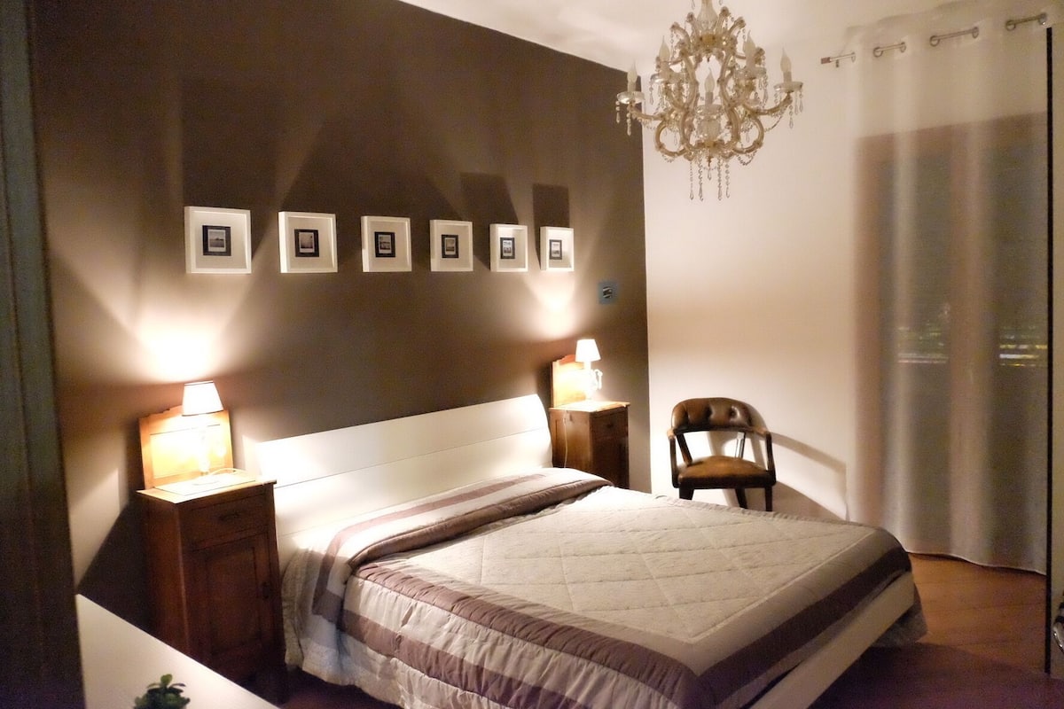 Un dormitorio ordenado con una cama, dos lámparas de noche, una silla, una araña y cuadros enmarcados en la pared.