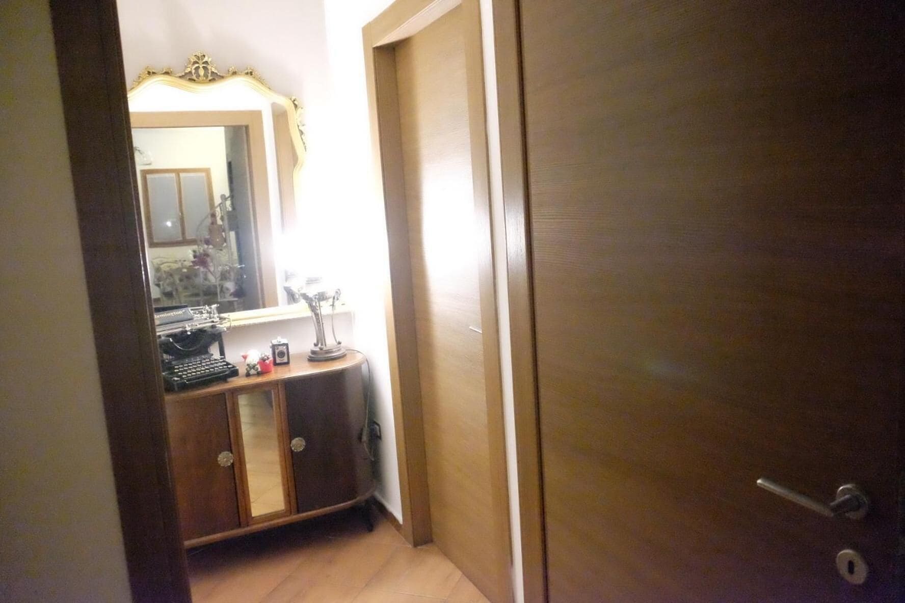 A narrow hallway with a wooden door on the right and a mirror reflecting a room with a desk and a chair.