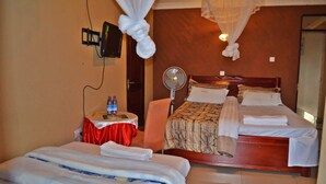A hotel room with two beds, a bedside table with water bottles, a TV mounted on the wall, and a standing fan. - Unik Hotel Limited (Kampala)