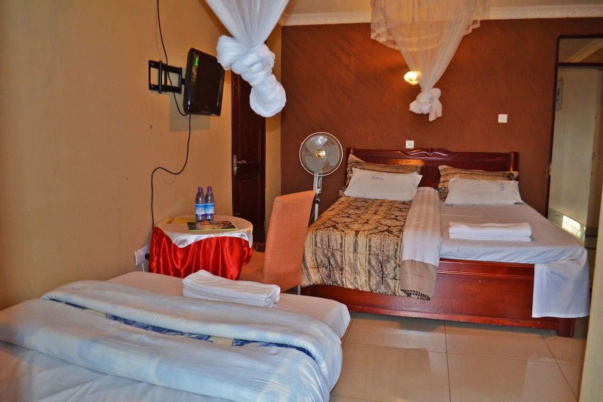A hotel room with two beds, a bedside table with a fan, a TV, and a mosquito net.