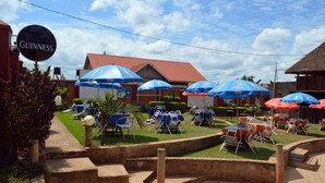 An outdoor seating area with tables and chairs under blue umbrellas, a sign for Guinness, and a building with a red roof. - Unik Hotel Limited (Kampala)