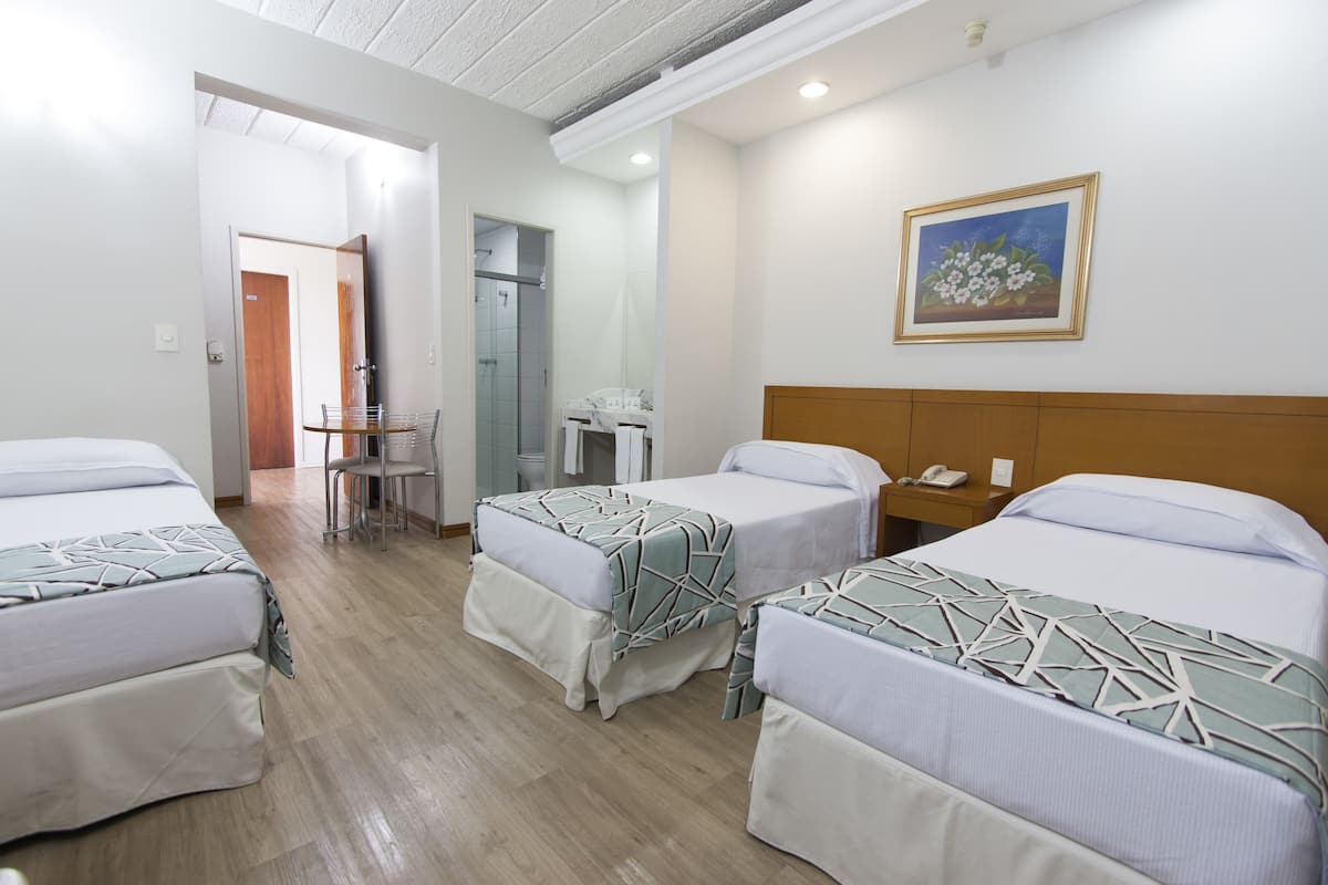 Une chambre d’hôtel avec trois lits, un bureau, une chaise et une salle de bain visible à travers une porte ouverte.