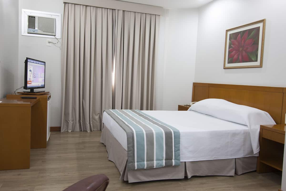 Une chambre d’hôtel avec un lit, un bureau, une chaise, une télévision, une fenêtre avec des rideaux et un tableau au mur.