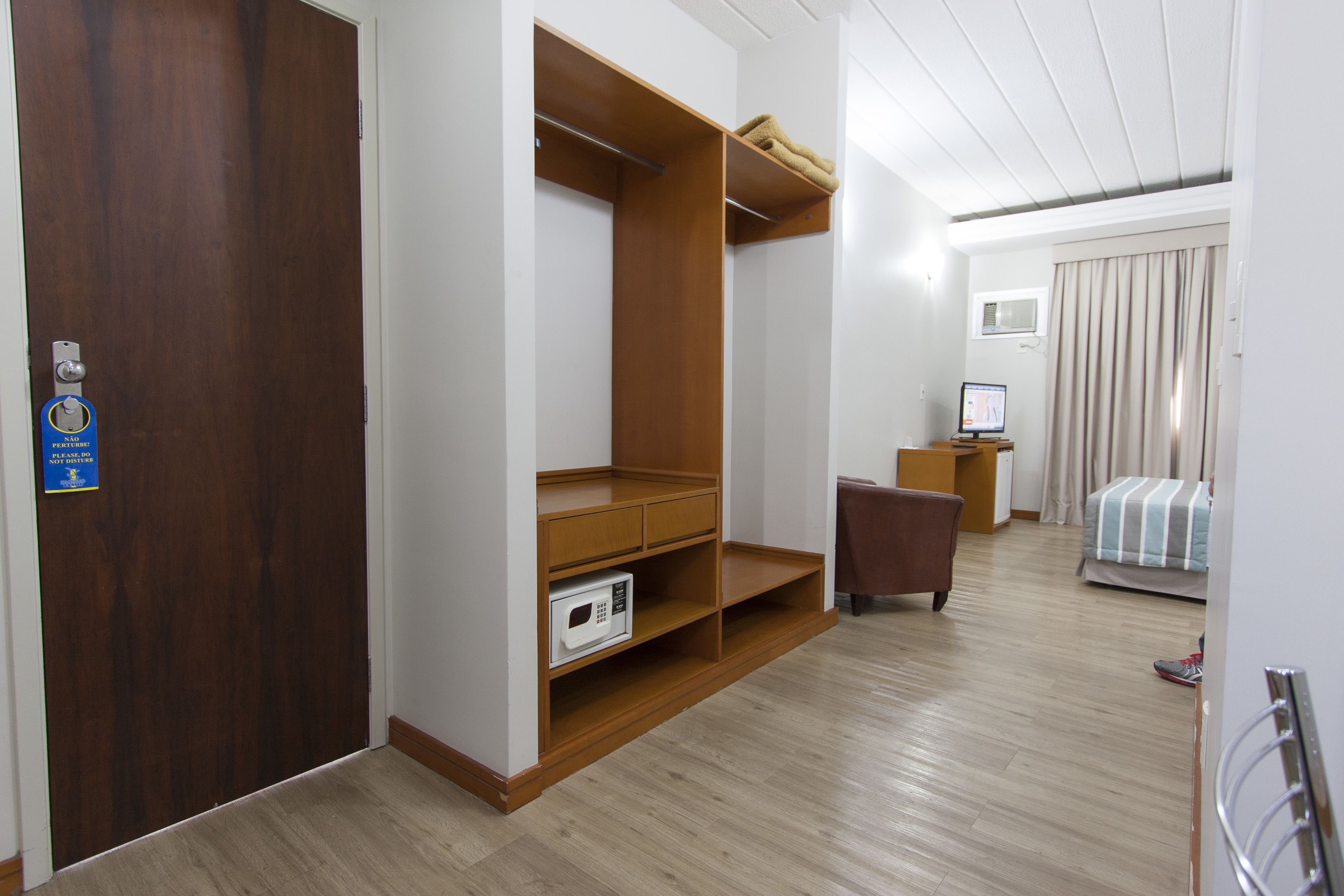 A hotel room with a wooden wardrobe, a small desk with a computer, a bed, and a window with curtains.