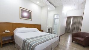 A hotel room with a large bed, a wooden headboard, a bedside table with a phone, a brown leather chair, a bathroom with a glass door, and a framed picture on the wall. - Di Giulio Hotel (São José dos Campos)