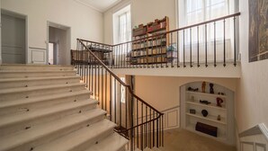 A grand staircase with a metal railing leading to a second floor, a large bookcase filled with books, and a fireplace with decorative items. - La Maison de Maître (Fontenay-le-Comte)