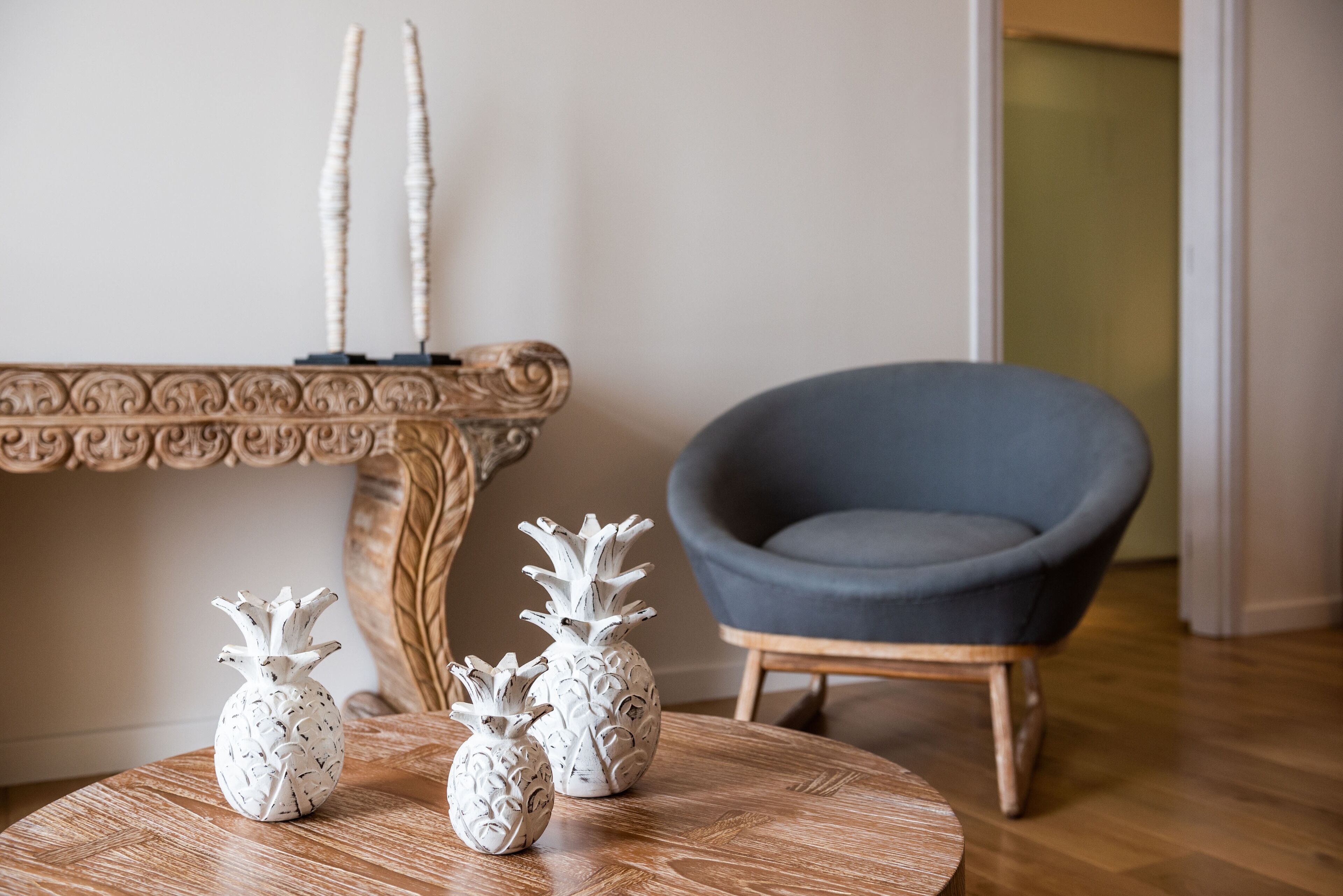Ein Zimmer mit einem runden Holztisch, drei weißen, ananasförmigen Vasen, einem dunkelblauen Sessel und einer dekorativen Konsole mit zwei hohen, weißen Vasen.