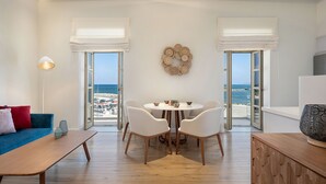 A modern living room with a dining area, large windows with white curtains, a round table with chairs, and a view of the sea. - Avra Apartments Kolymbari Harbour (Platanias)