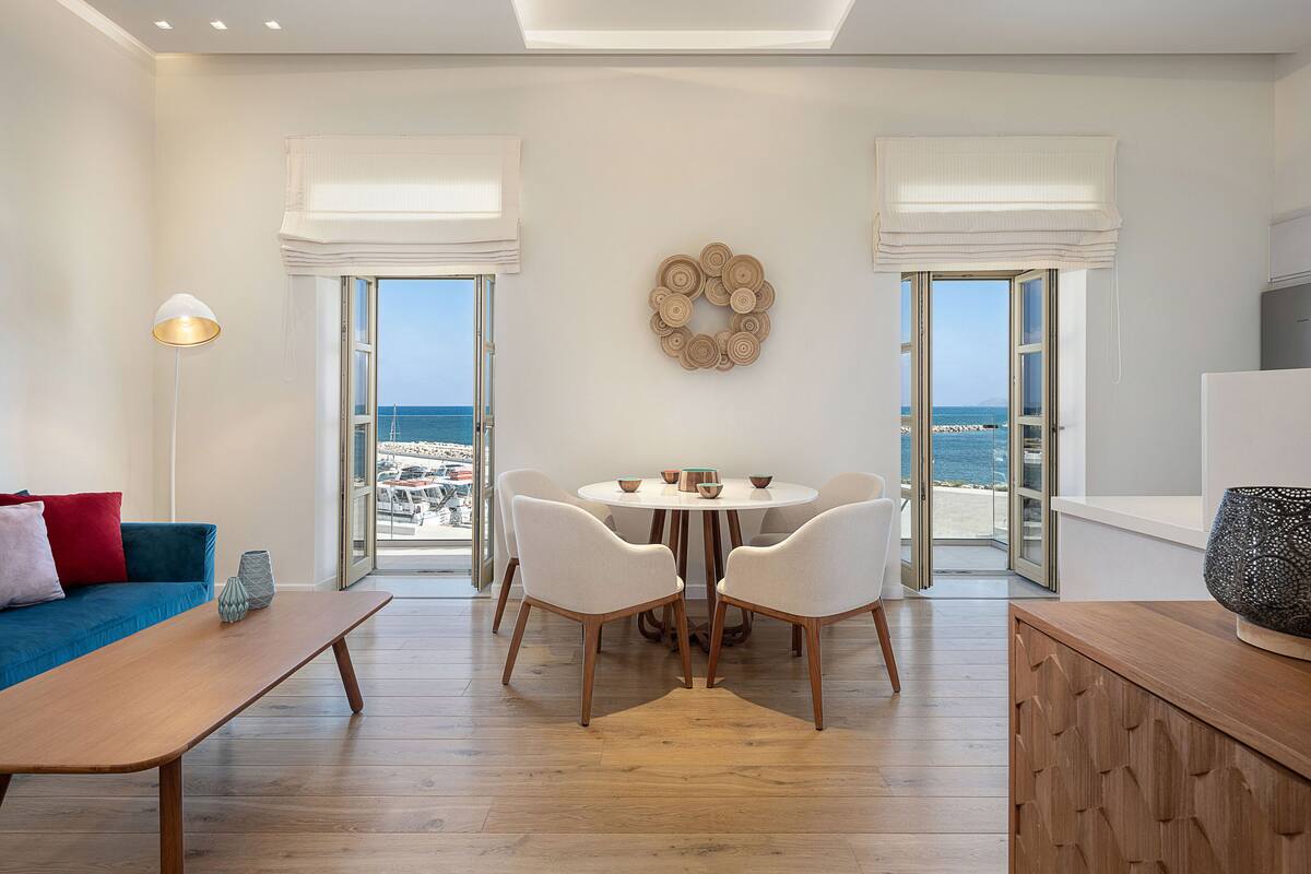 A modern living room with a dining area, large windows with white curtains, a round table with chairs, and a view of the sea.