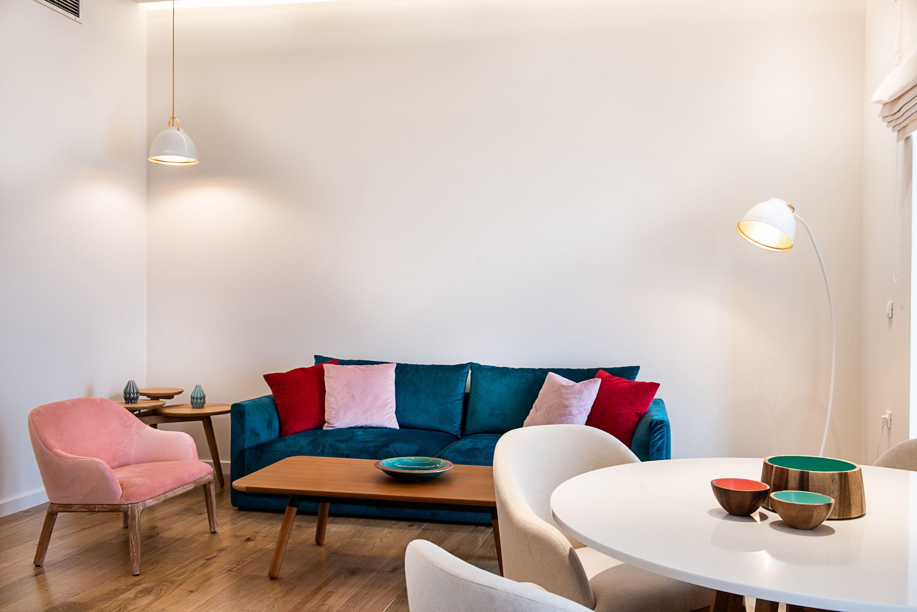 A modern living room with a blue sofa, a pink armchair, a wooden coffee table, and a round dining table with three bowls.